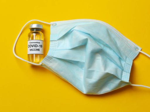 A facemask and a COVID-19 vaccine vile on a yellow background