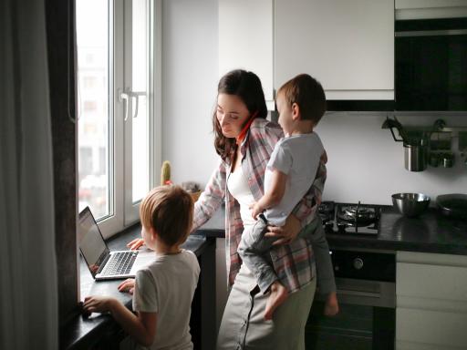A parent is holding a child and working on their laptop in their kitchen.