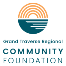 Logo for Grand Traverse Regional Community Foundation with a circle above the text that has two colors - green on the bottom half and orange on the top half. The orange appears to be the sun represented by an all orange rainbow shape and the land represented by the bottom half of the circle in dark green 