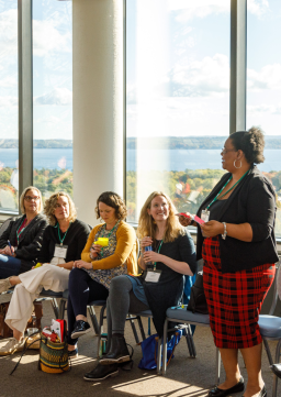 An woman speaks during a workshop at a conference with a room full of people who are sat in a circle, listening intently and you can see the fall leaves changing color through the windows behind the group and a view of the water beyond the leaves