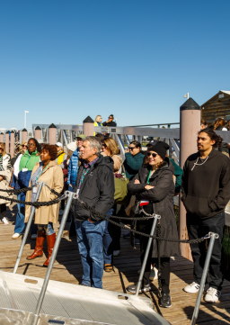 A group of conference attendees listening to someone off camera while they stand on a dock waiting to board a sailing boat on the West Traverse Bay
