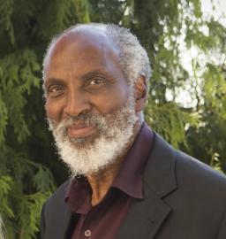 A professional headshot of john powell, wearing a black suit jacket and dark purple buttondown shirt, smiling at the camera