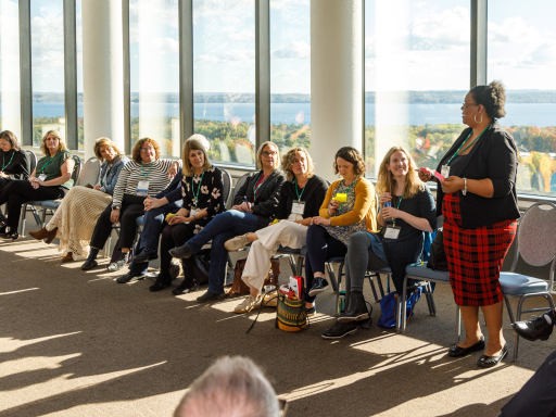 An woman speaks during a workshop at a conference with a room full of people who are sat in a circle, listening intently and you can see the fall leaves changing color through the windows behind the group and a view of the water beyond the leaves