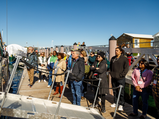 A group of conference attendees listening to someone off camera while they stand on a dock waiting to board a sailing boat on the West Traverse Bay
