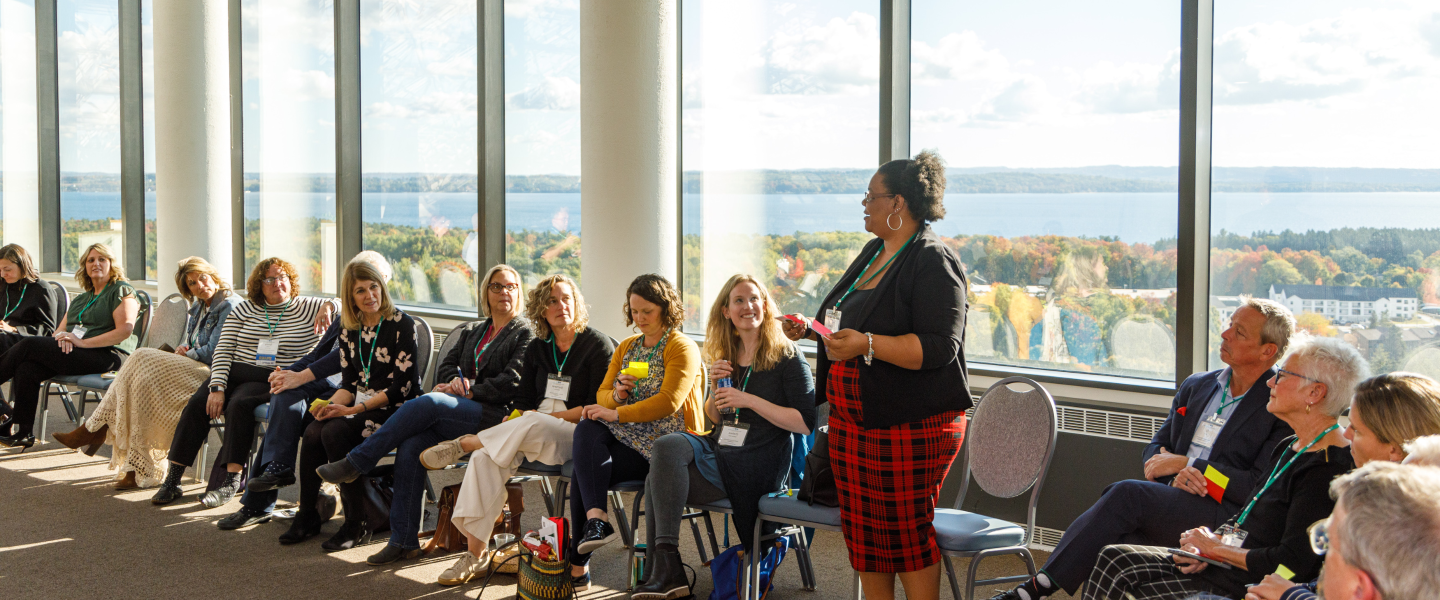 An woman speaks during a workshop at a conference with a room full of people who are sat in a circle, listening intently and you can see the fall leaves changing color through the windows behind the group and a view of the water beyond the leaves