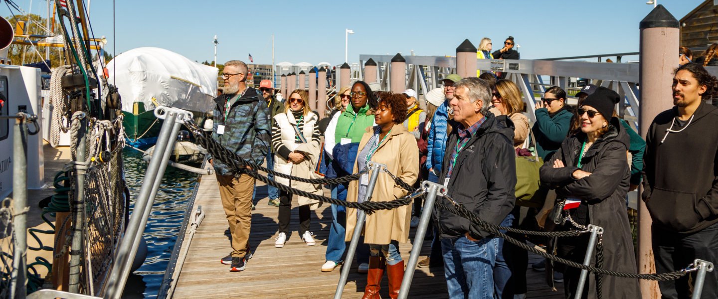 A group of conference attendees listening to someone off camera while they stand on a dock waiting to board a sailing boat on the West Traverse Bay