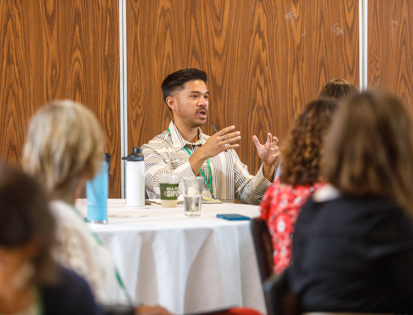 An individual sitting in a room full of tables during a program with other members, discussing something and engaging with other participants