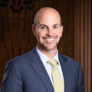 Headshot for Ridgeway White, wearing a dark suit and light shirt and yellow tie, smiling at the camera