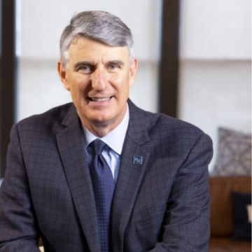 Headshot for Dave Egner, wearing a black suit with collared shirt and blue tie, smiling at the camera