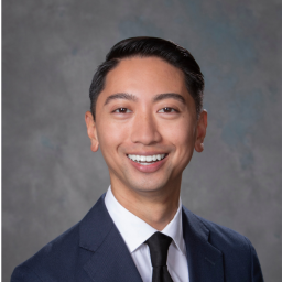 Headshot for JD Lesada, who is wearing a dark blue suit, white shirt and dark blue tie, and smiling at the camera