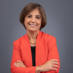 Headshot for Maha Freij, smiling at the camera and wearing a burnt orange suit jacket with arms crossed in front of a grey background