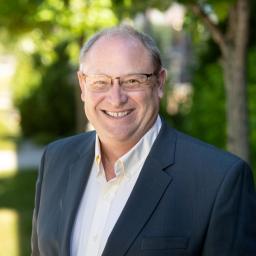 Randy is smiling, standing outside wearing a suit and a light colored shirt.